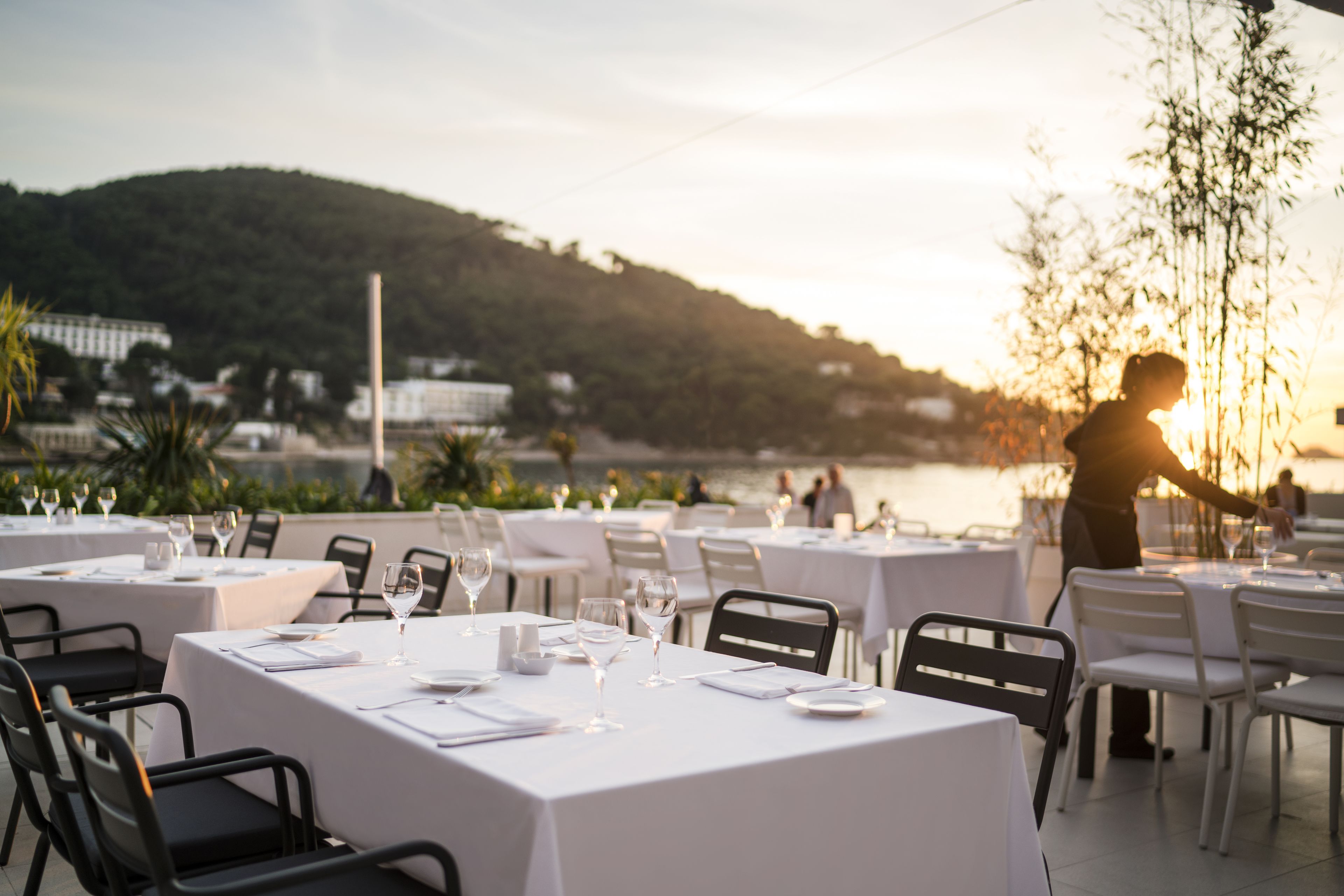 A restaurant with tables set for dining, overlooking a body of water and a hill.