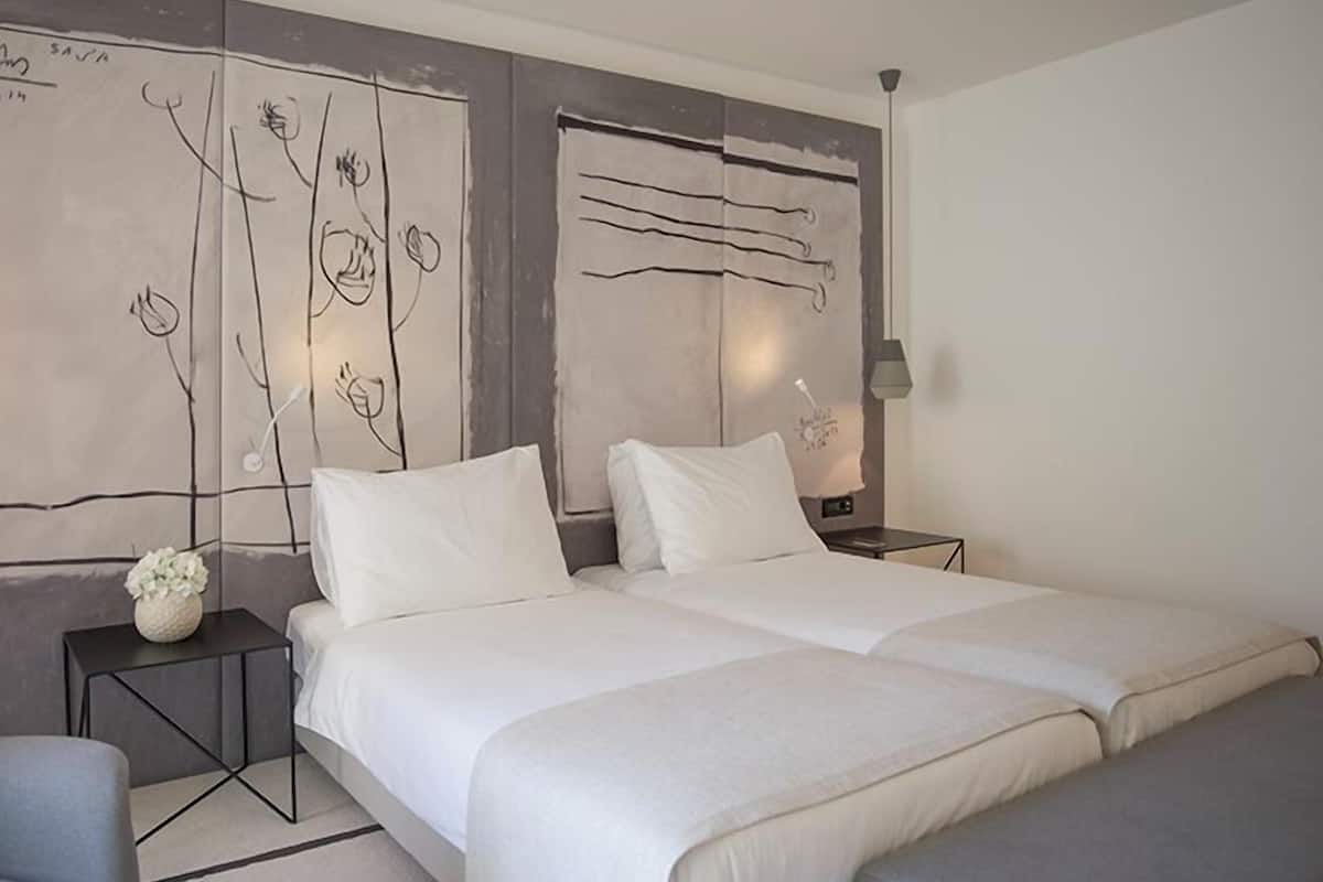 Una habitación de hotel moderna con dos camas, una mesita de noche con lámpara y una pared decorativa con arte abstracto.