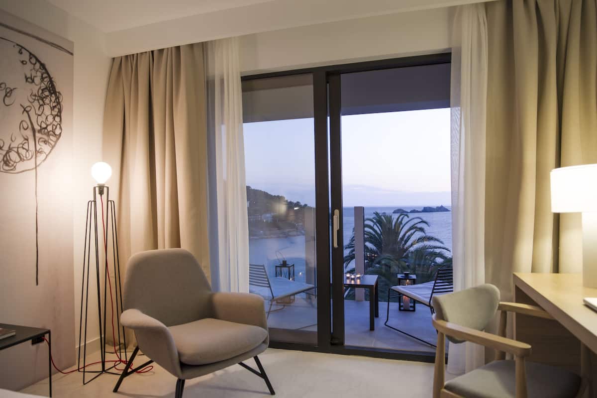 Habitación de hotel con un gran ventanal corredero que da a un balcón con vistas al mar y palmeras. Hay dos sillas y una mesita en el balcón.
