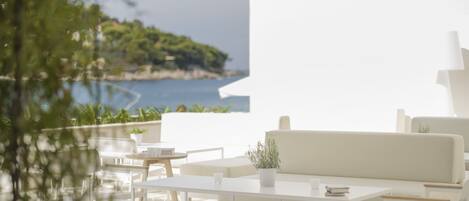 A modern outdoor seating area with white furniture, a view of the sea, and a white building in the background.