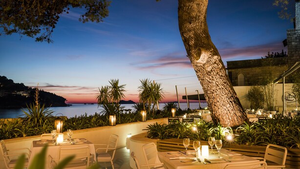 An outdoor dining area with tables set for two, overlooking a body of water at sunset. - Hotel Kompas (Dubrovnik)