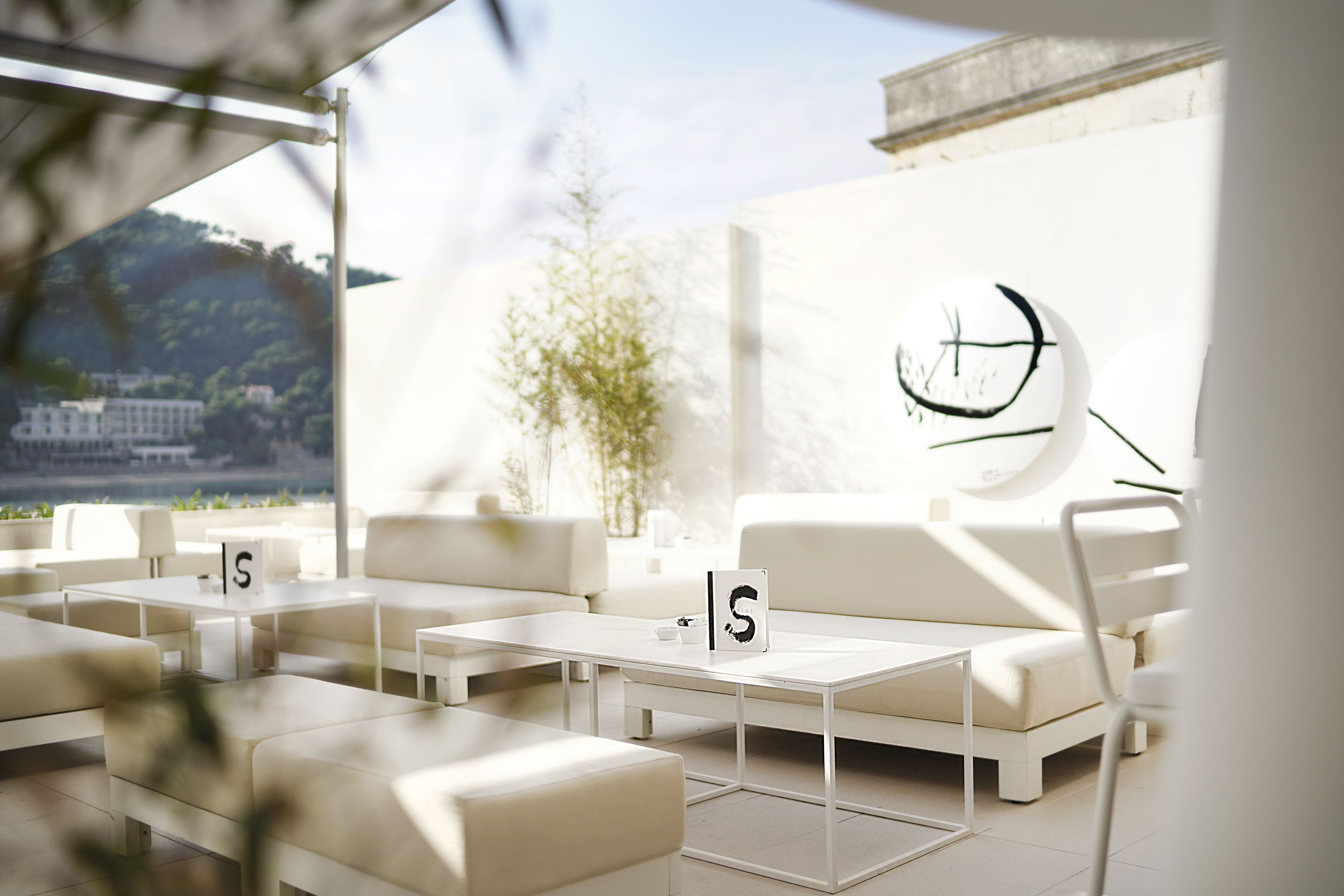 Un área de estar exterior moderna con mobiliario blanco, diseño minimalista y vista a un edificio y follaje al fondo.
