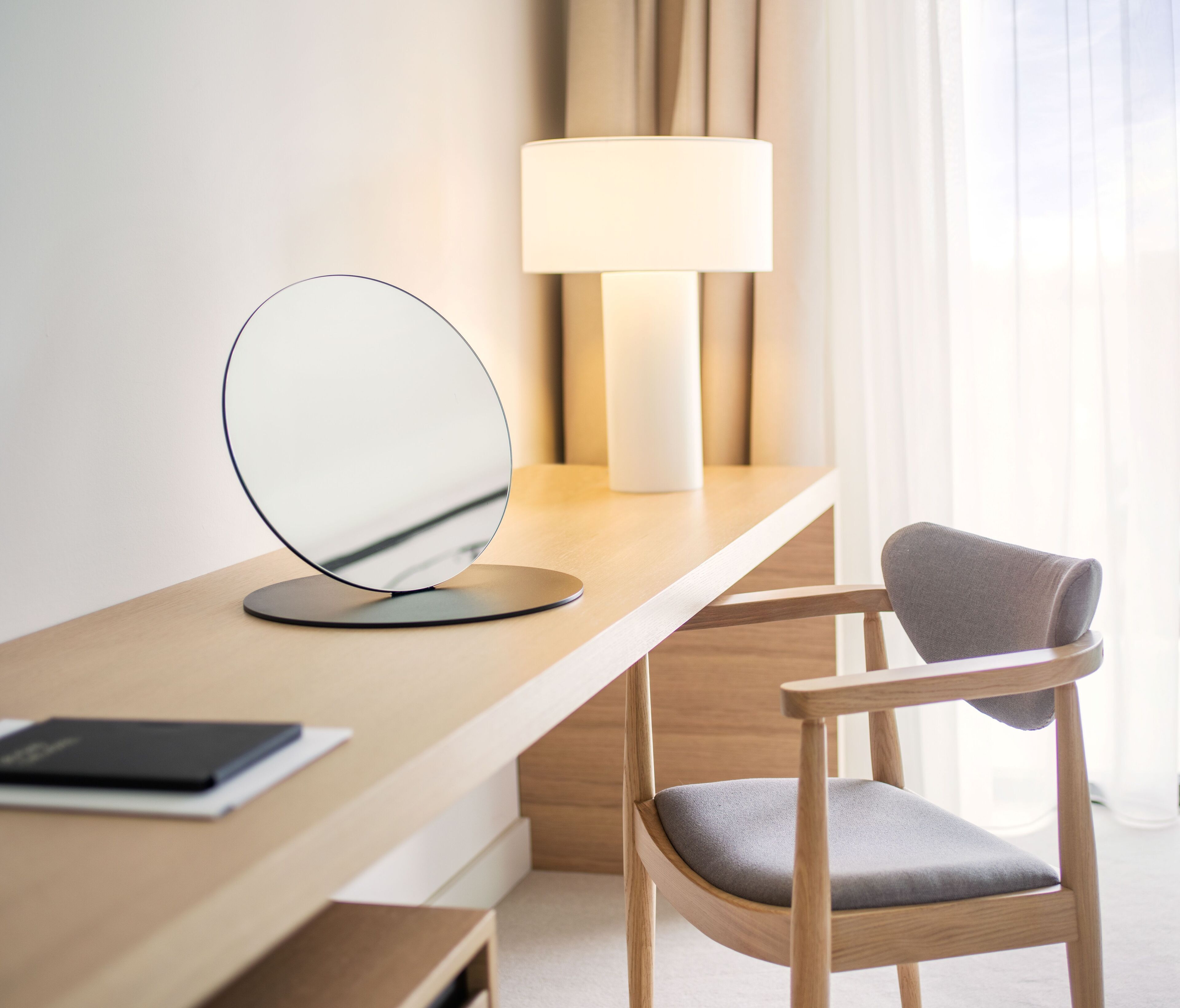 A modern hotel room with a wooden desk, a round mirror, a lamp, and a chair.
