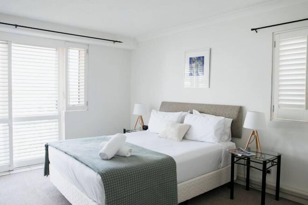 A hotel room with a bed, two bedside tables, a window with blinds, and a picture on the wall.