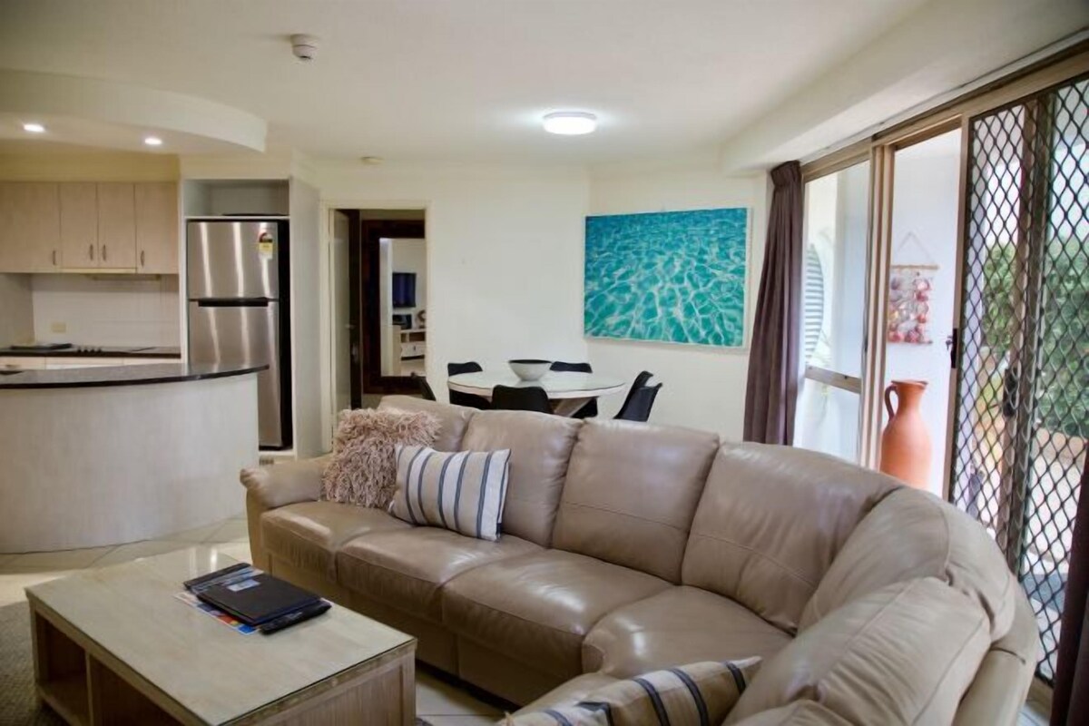 A modern living room with a beige leather sofa, a coffee table, and a dining area with a white table and chairs.