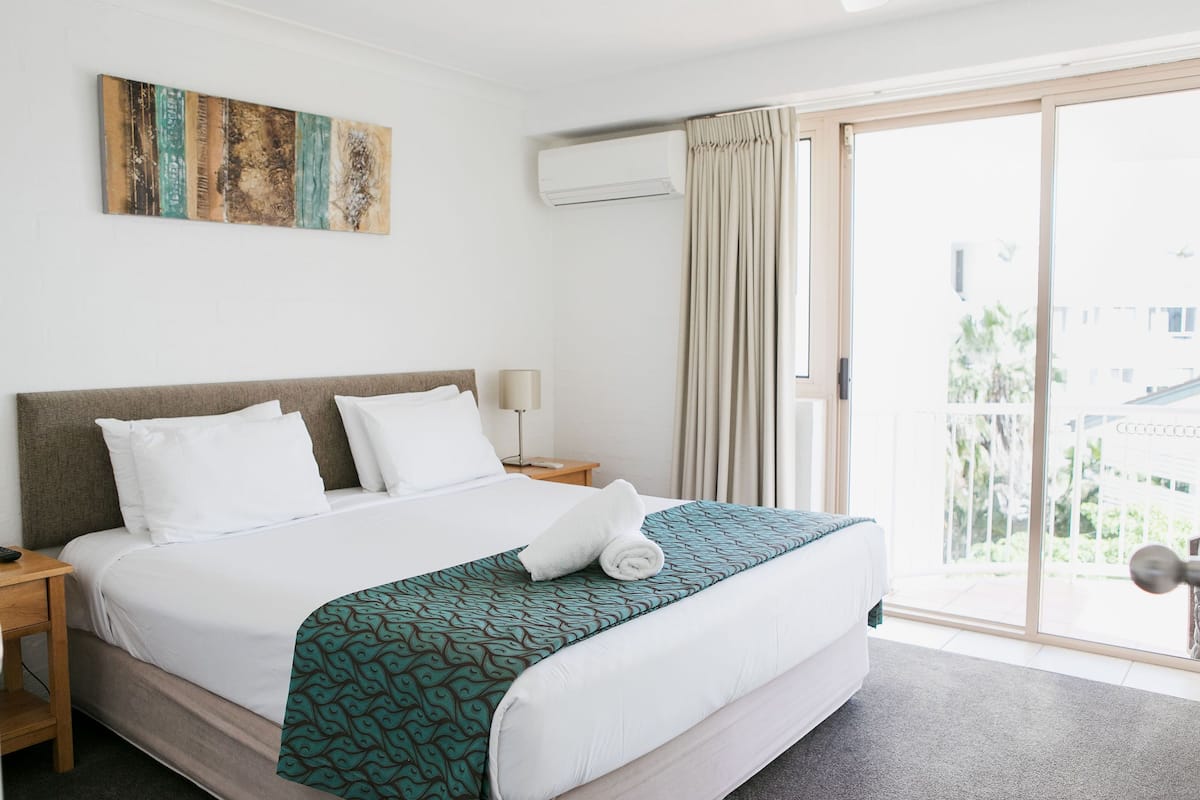 A hotel room with a bed, a nightstand, a wall-mounted air conditioner, and a view of the outside through a sliding glass door.