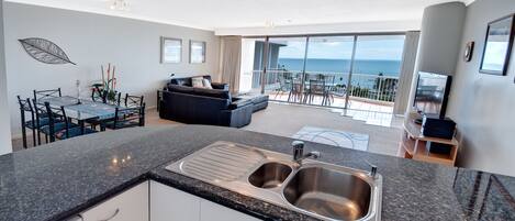 Cocina moderna con pileta doble, comedor con mesa y sillas, un sofá y un balcón con vista al océano.