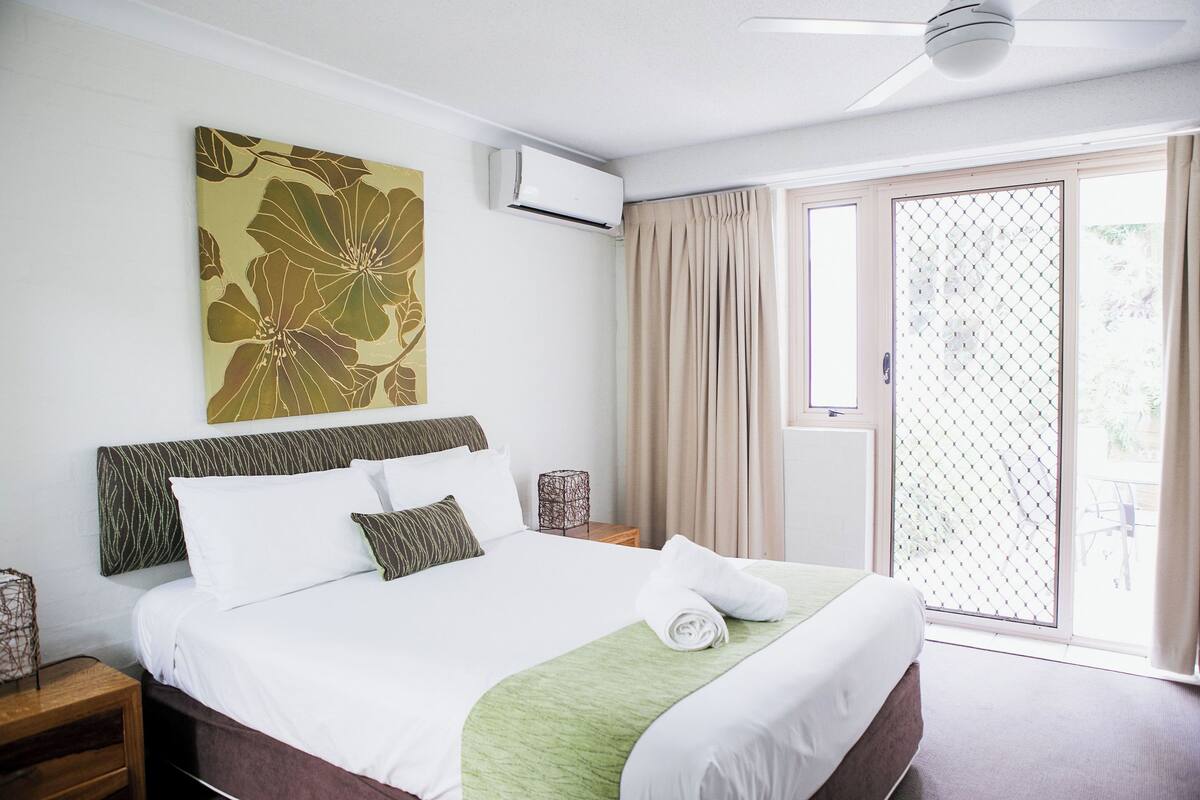 A hotel room with a bed, an air conditioner, a window with curtains, and a view of the outdoors.