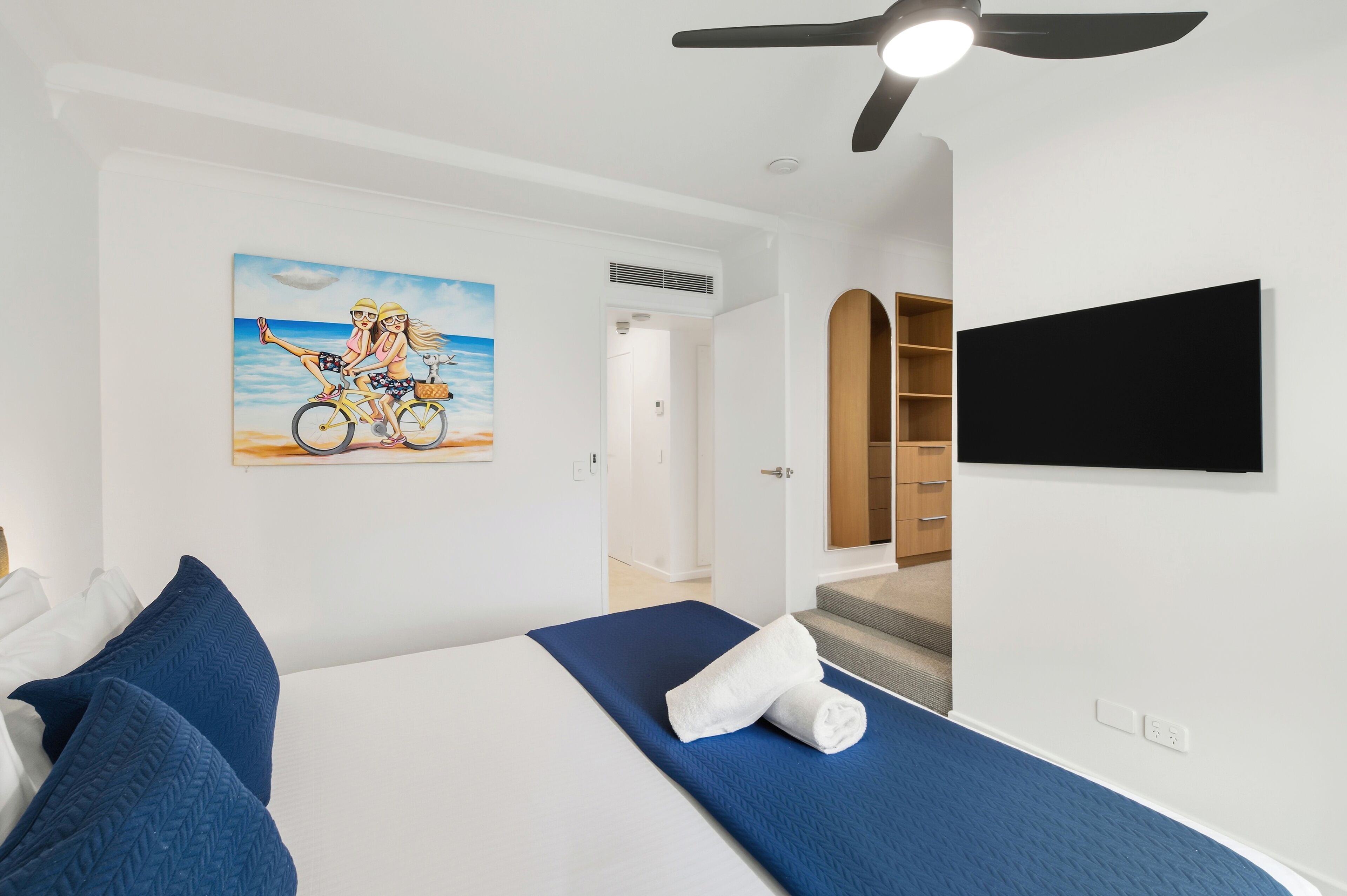 A hotel room with a bed, a painting of two people on a bicycle, a ceiling fan, and a TV mounted on the wall.