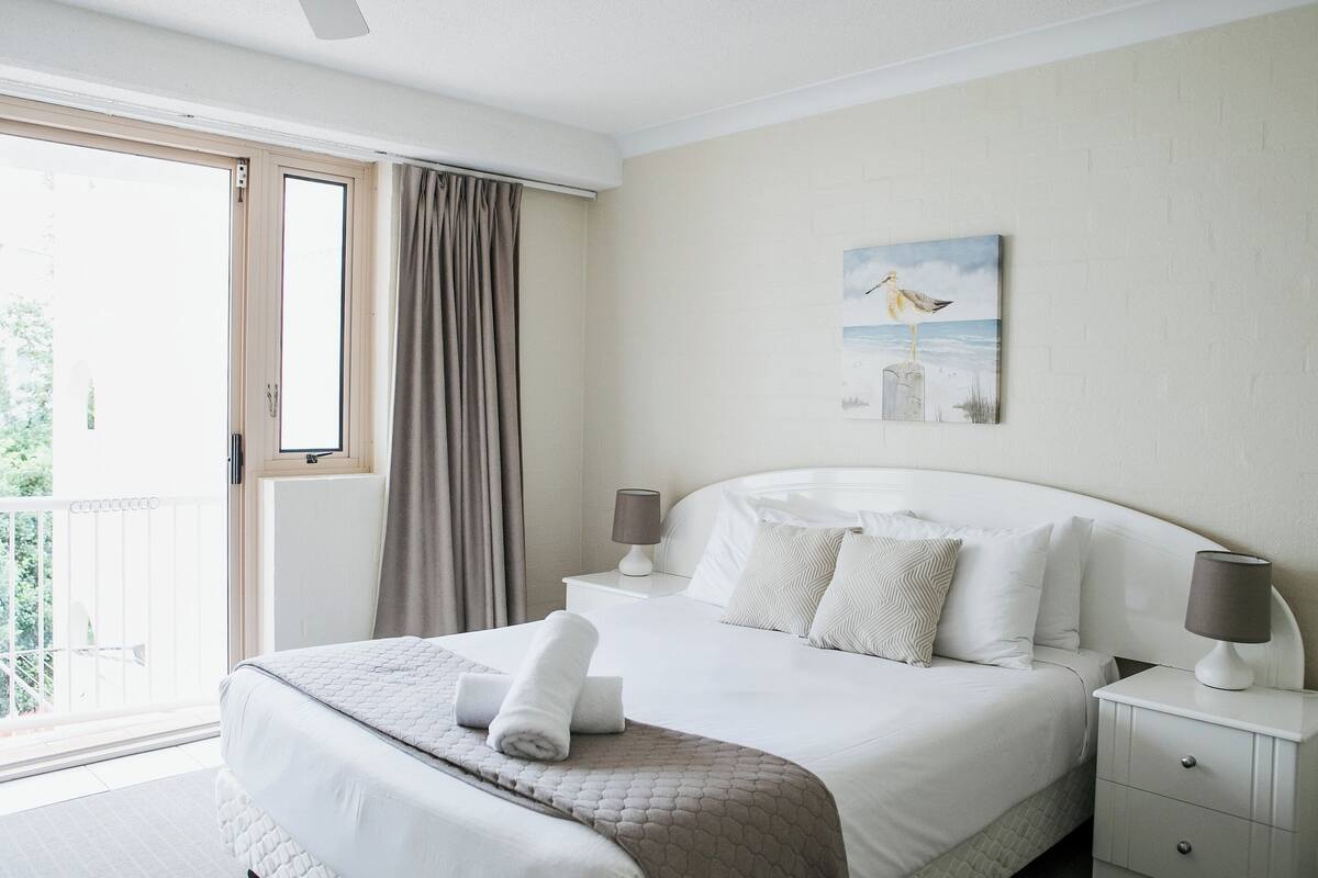 A hotel room with a bed, a nightstand, a lamp, a painting, and a view of the outdoors.