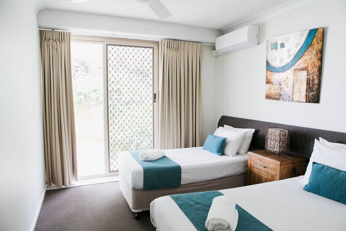 A hotel room with two beds, a wooden nightstand, a wall-mounted air conditioner, and a sliding glass door leading to an outdoor area.