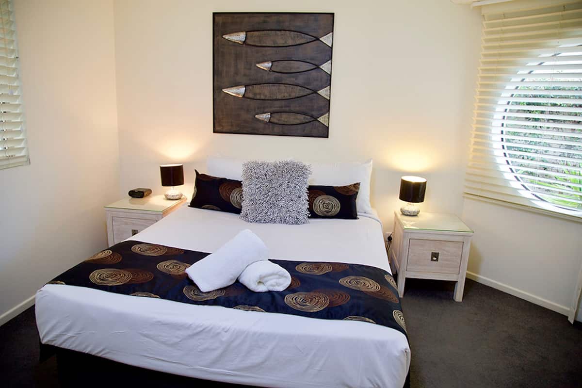 A neatly made bed with a patterned bedspread, two bedside lamps, a decorative wall piece, and a window with blinds.