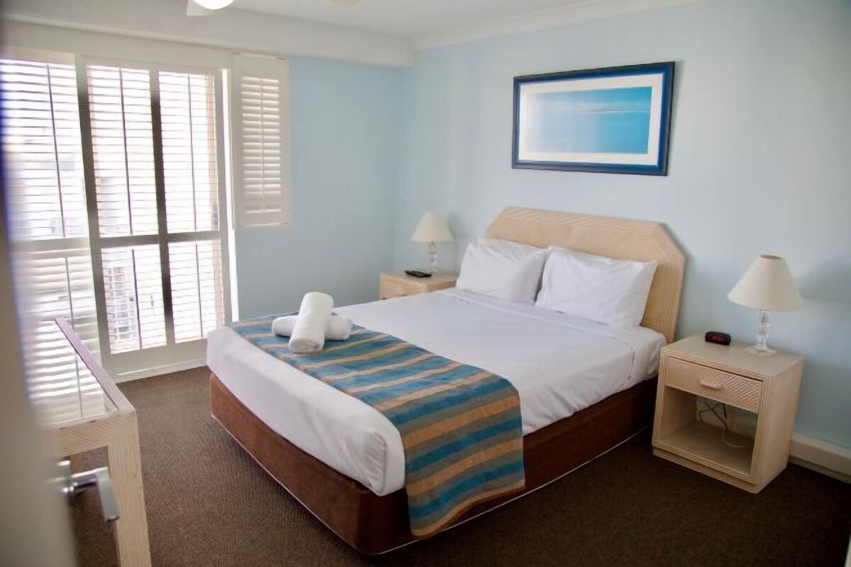 A hotel room with a bed, two bedside tables with lamps, a window with blinds, and a framed picture on the wall.