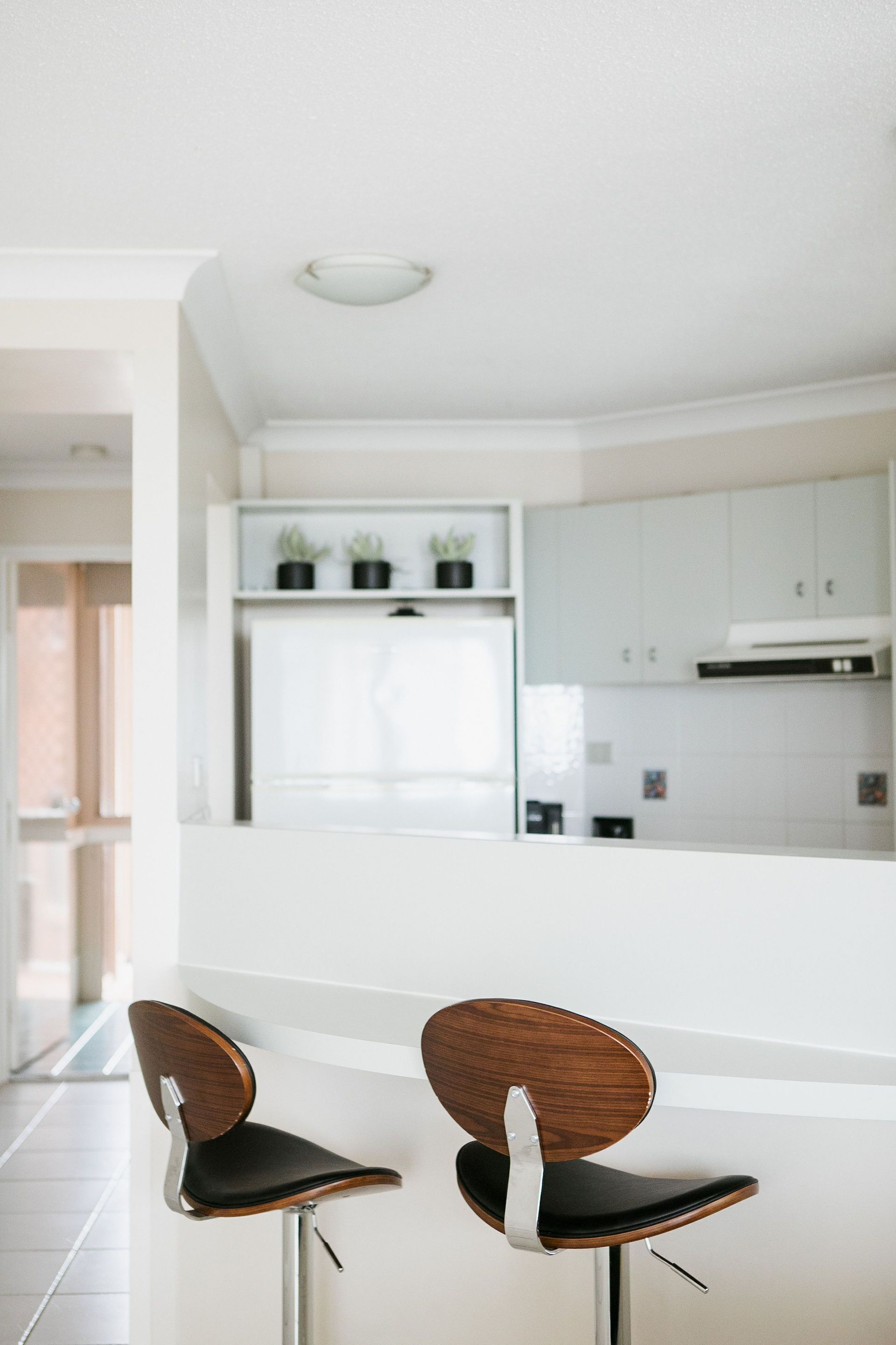 A modern kitchen with white cabinets and a built-in stove. There are two wooden chairs with metal legs and black cushions.