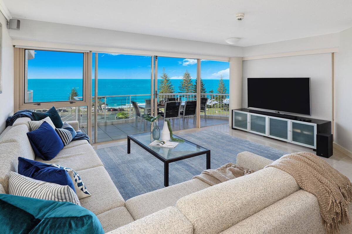 A modern living room with a large sofa, a coffee table, and a flat-screen TV. The room has large windows offering a view of the ocean and trees.