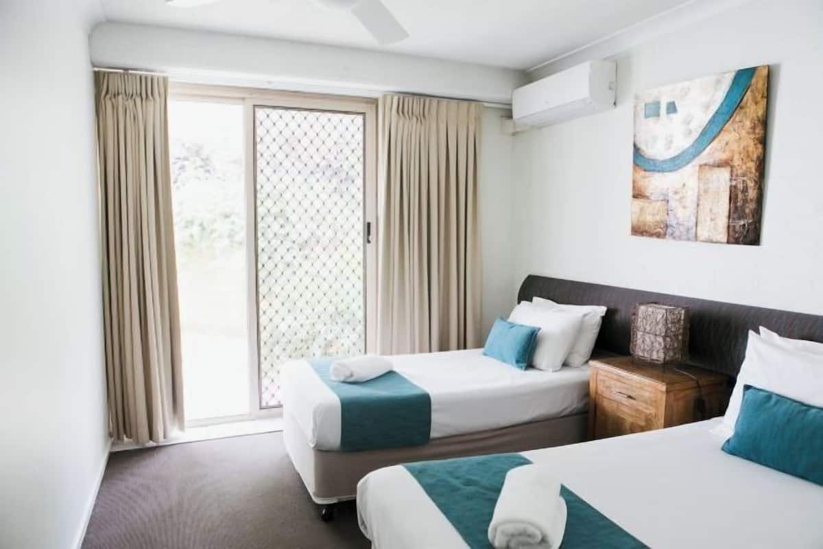 A hotel room with two beds, a window with curtains, a wall-mounted air conditioner, and a painting.