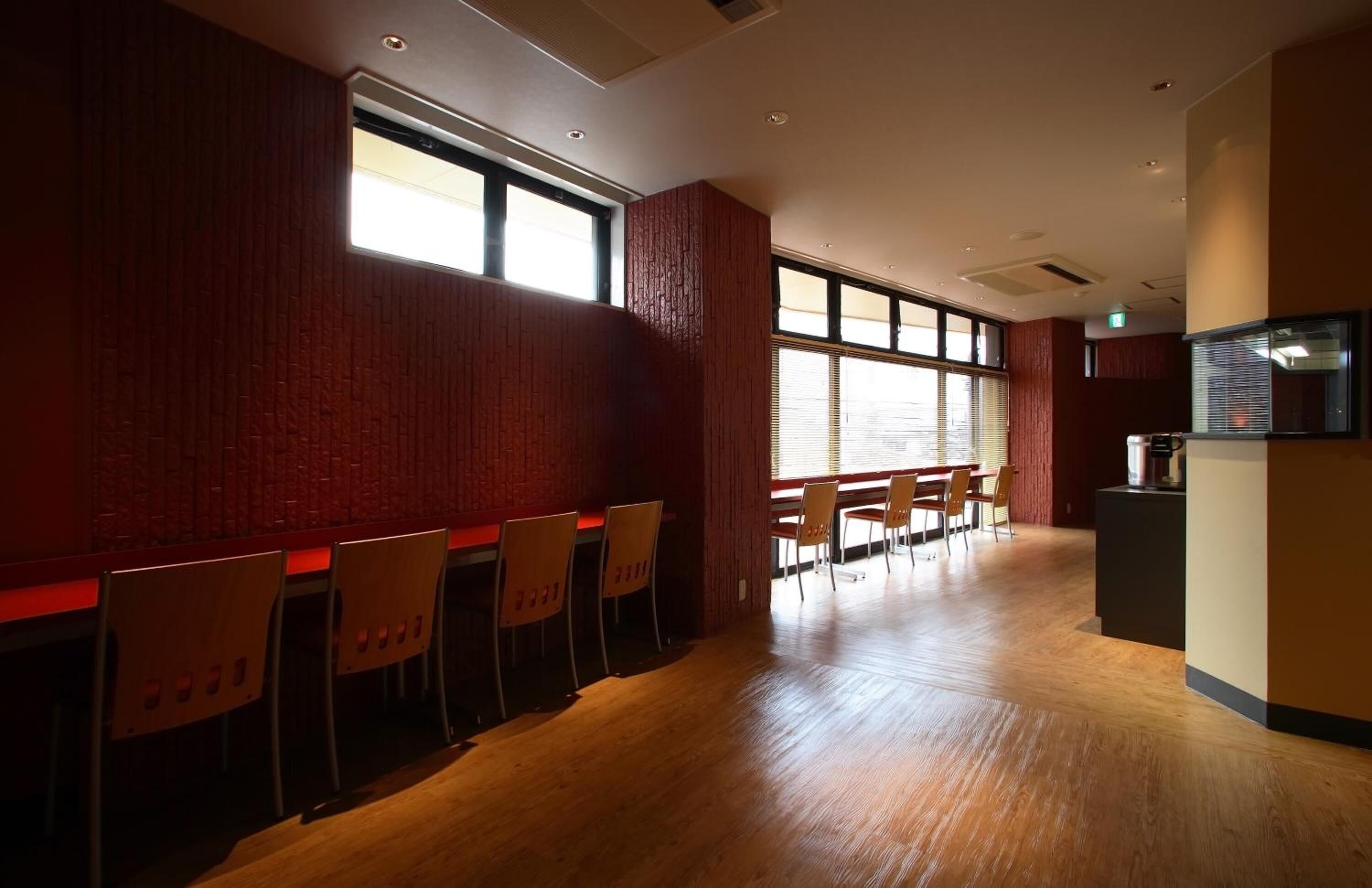 A spacious room with wooden flooring, red brick walls, and large windows. There are several wooden chairs and tables arranged for dining.
