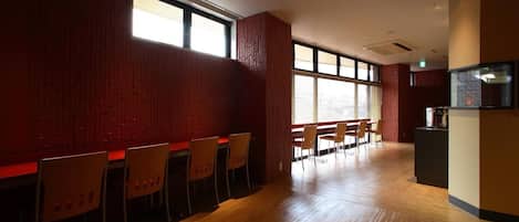 A spacious room with wooden flooring, red brick walls, and large windows. There are several wooden chairs and tables arranged for dining.