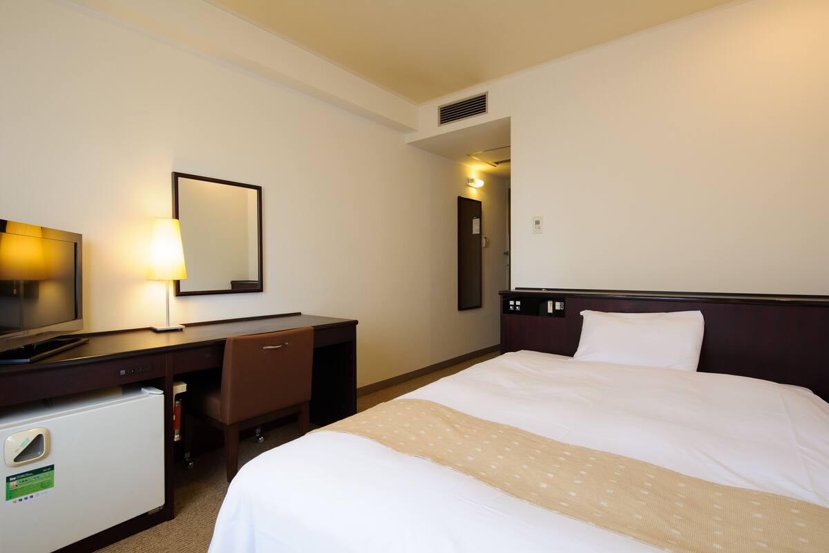 Habitación de hotel con cama, escritorio con lámpara, refrigerador pequeño y espejo en la pared.