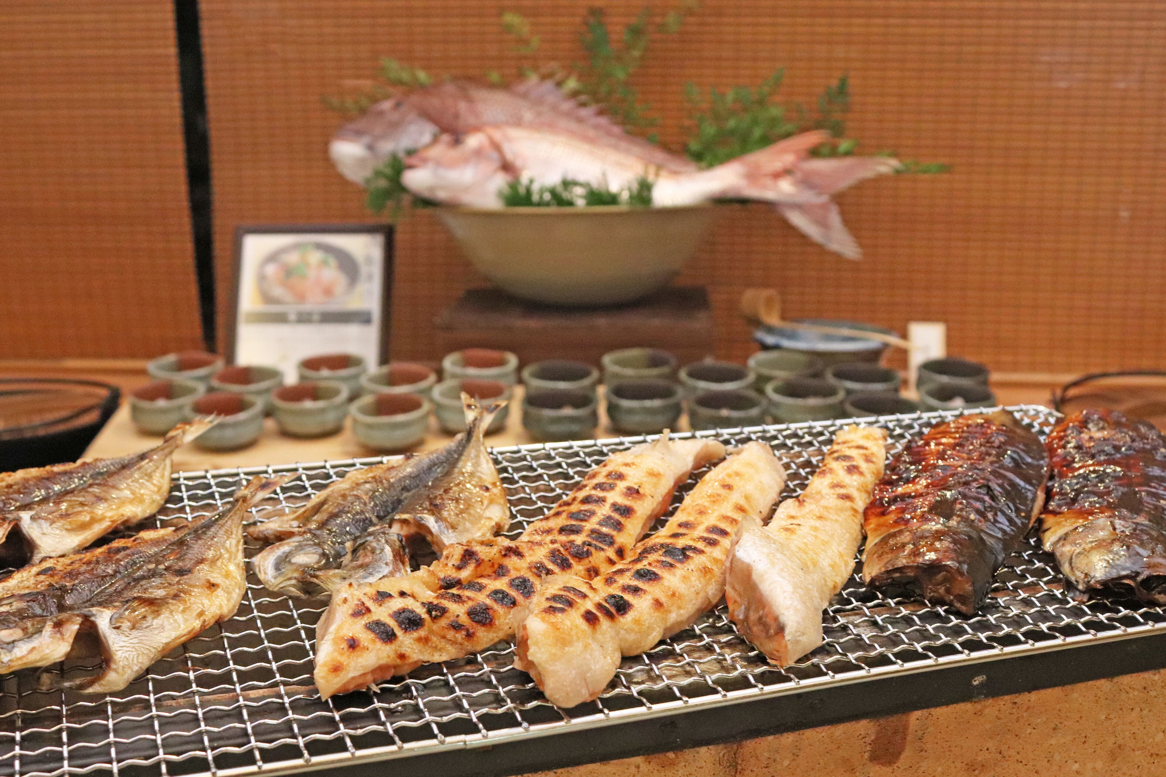 A variety of grilled fish and seafood on a grill, with small bowls of sauces in the background.