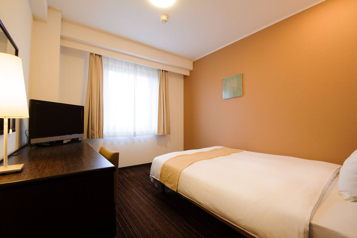Una habitación de hotel con cama, escritorio con televisor y ventana con cortinas.