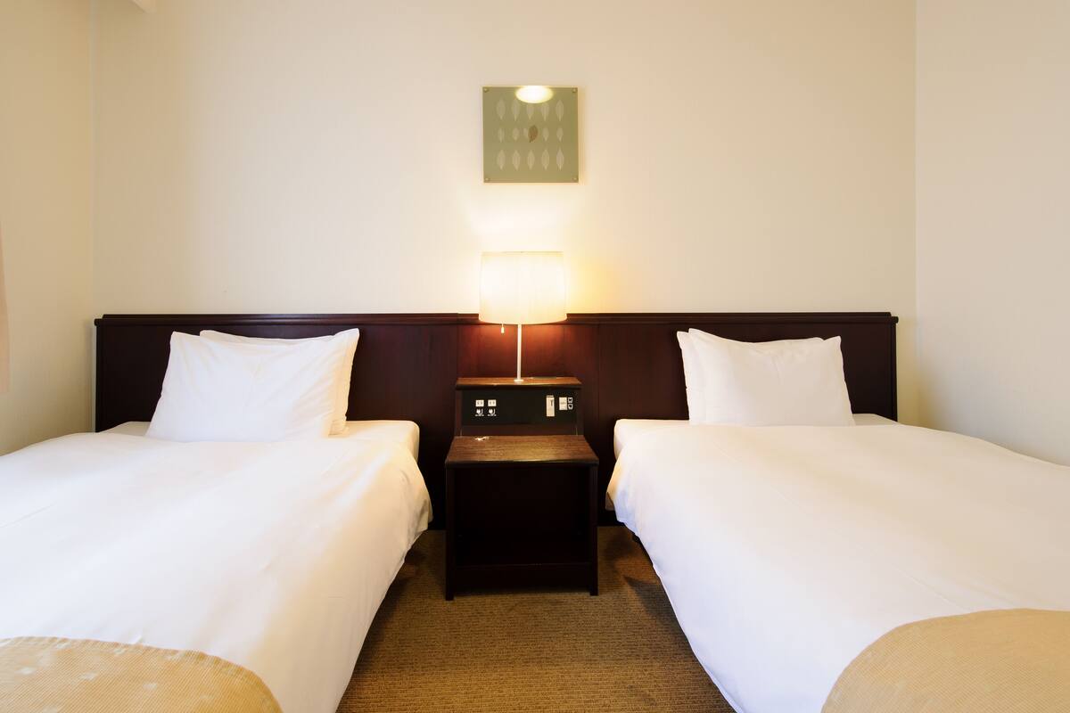 Habitación de hotel con dos camas, una mesita de noche con lámpara y una lámpara de pared.
