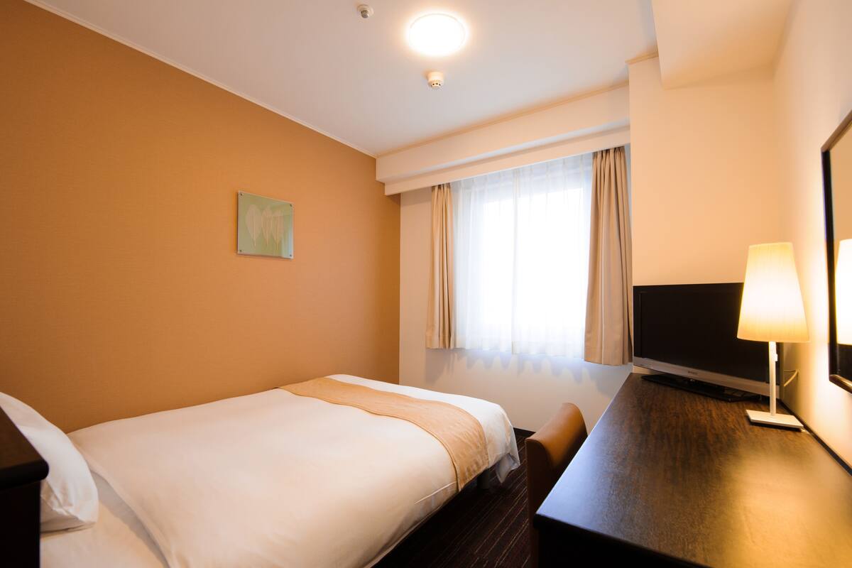 Una habitación de hotel con cama, escritorio con televisor, ventana con cortinas y un cuadro en la pared.