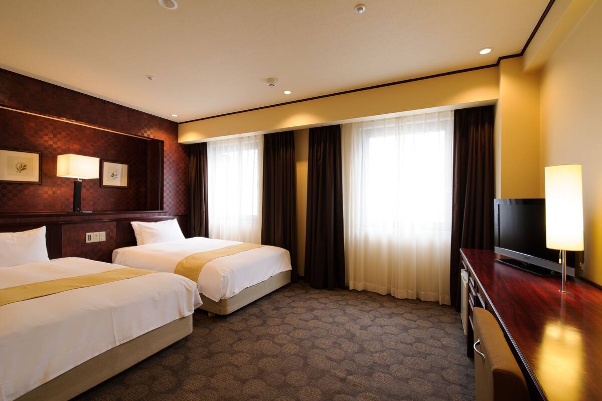 Habitación de hotel con dos camas, televisión, escritorio y ventana con cortinas.