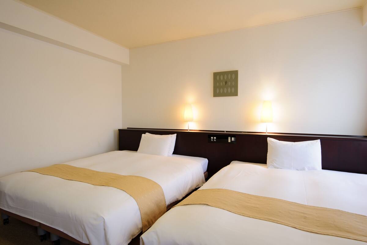 Habitación de hotel con dos camas individuales, cada una con una manta color beige y ropa de cama blanca. Dos lámparas de pared proporcionan iluminación.