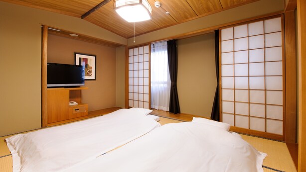 Una habitación tradicional japonesa con suelo de tatami, una cama con ropa blanca, un televisor sobre un mueble de madera y puertas correderas shoji.