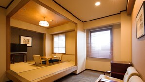 Una habitación con techo bajo, suelo de esteras de tatami, una cama, un escritorio, una silla, un televisor y una ventana con persianas.