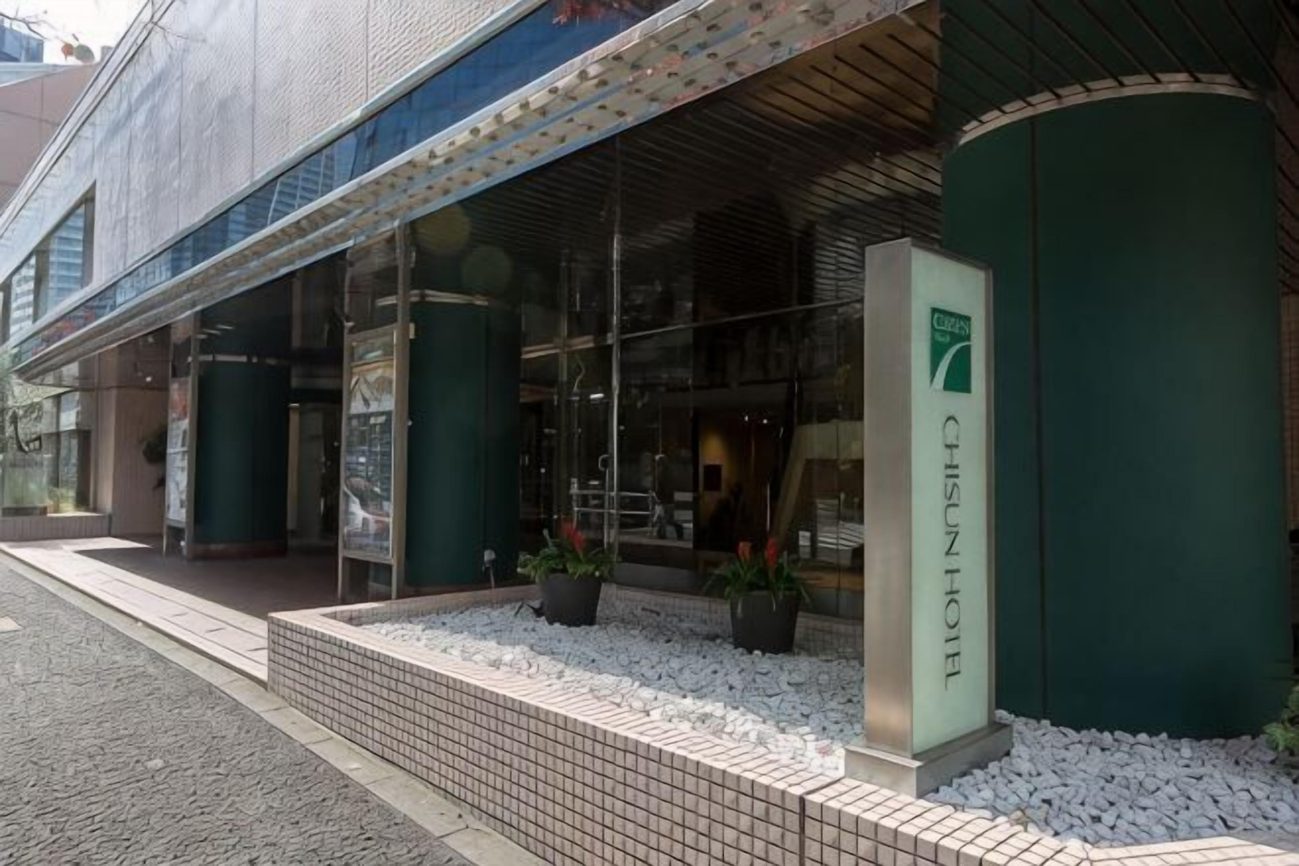 A hotel entrance with a glass facade, a signboard, and potted plants.