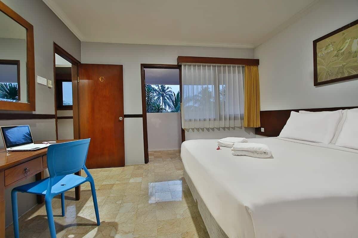 A hotel room with a bed, a desk with a laptop, a chair, a door leading to a balcony, and a window with curtains.