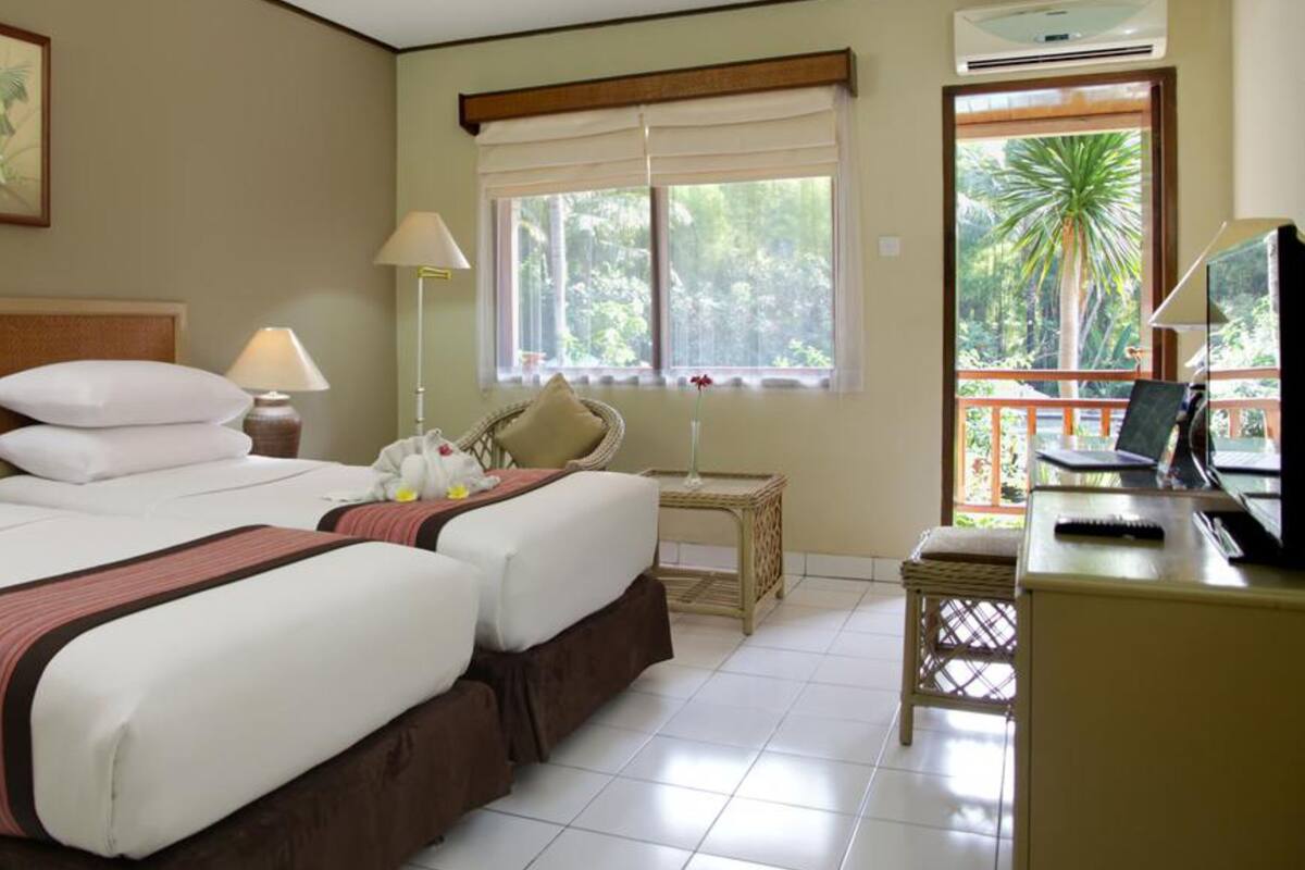Habitación de hotel con dos camas, un escritorio con una computadora portátil, una silla, una ventana que ofrece vista a palmeras y un cuadro en la pared.