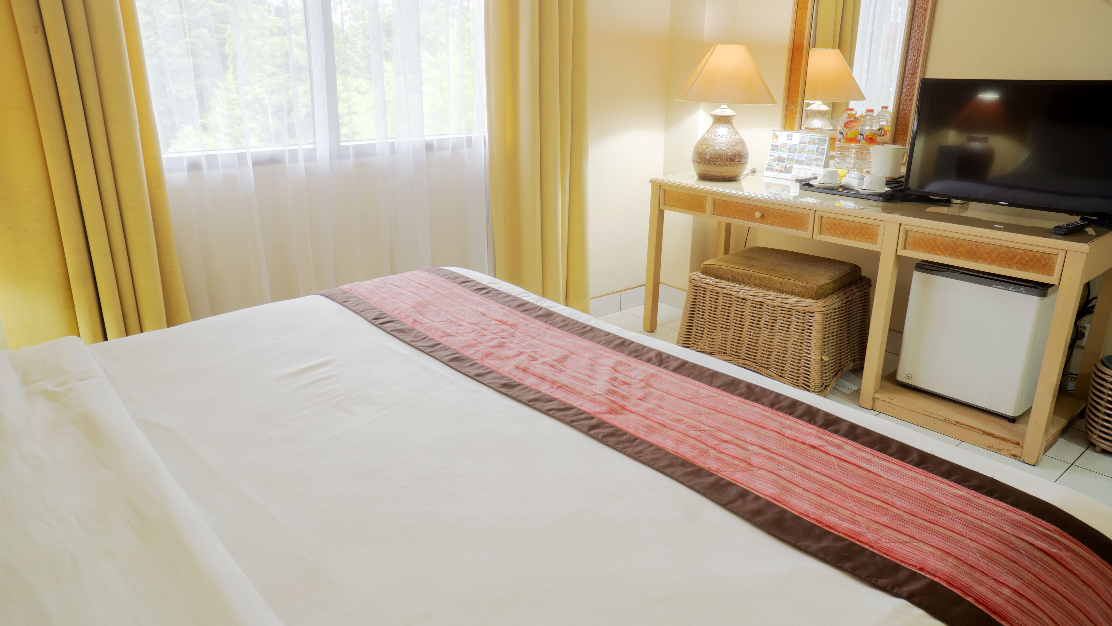 A hotel room with a bed, a desk with a television, a small refrigerator, and a wicker stool.