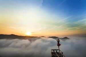 Una persona sobre una plataforma de madera, con vista al amanecer sobre un mar de nubes y colinas lejanas.