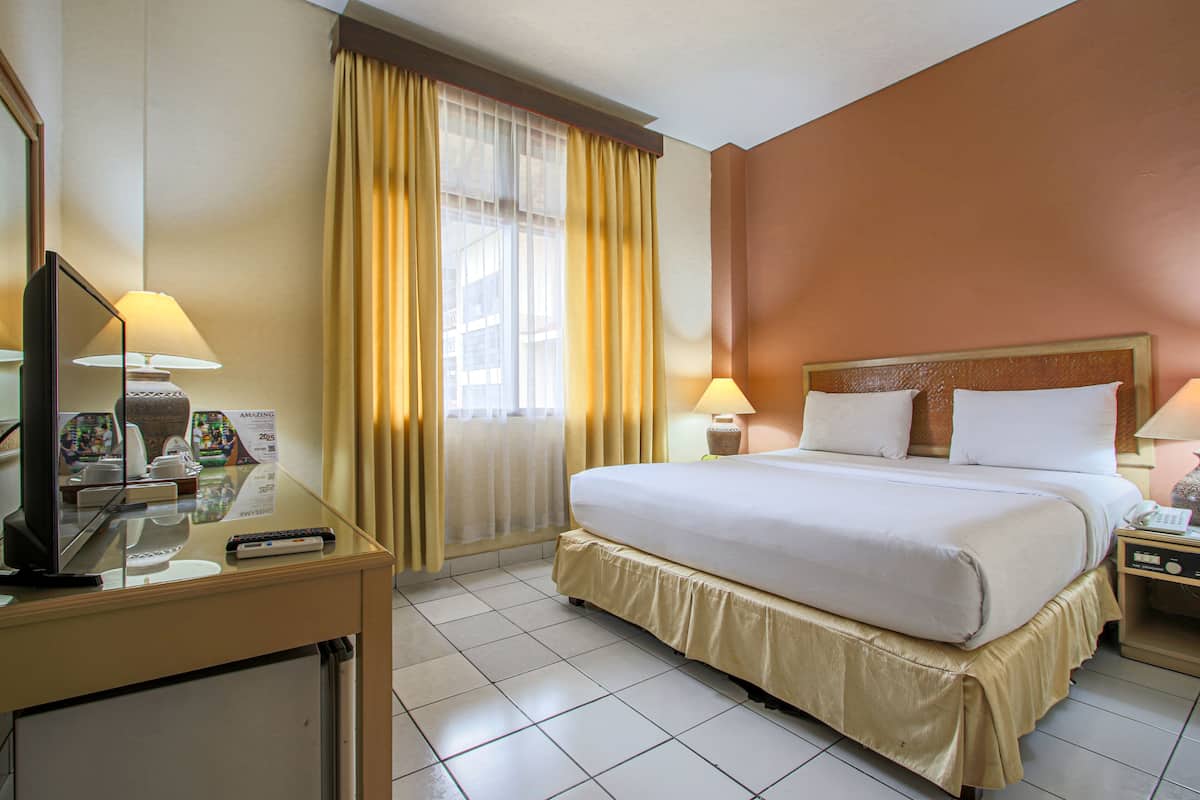Una habitación de hotel con cama, mesitas de noche, escritorio con lámpara, televisor y cortinas.