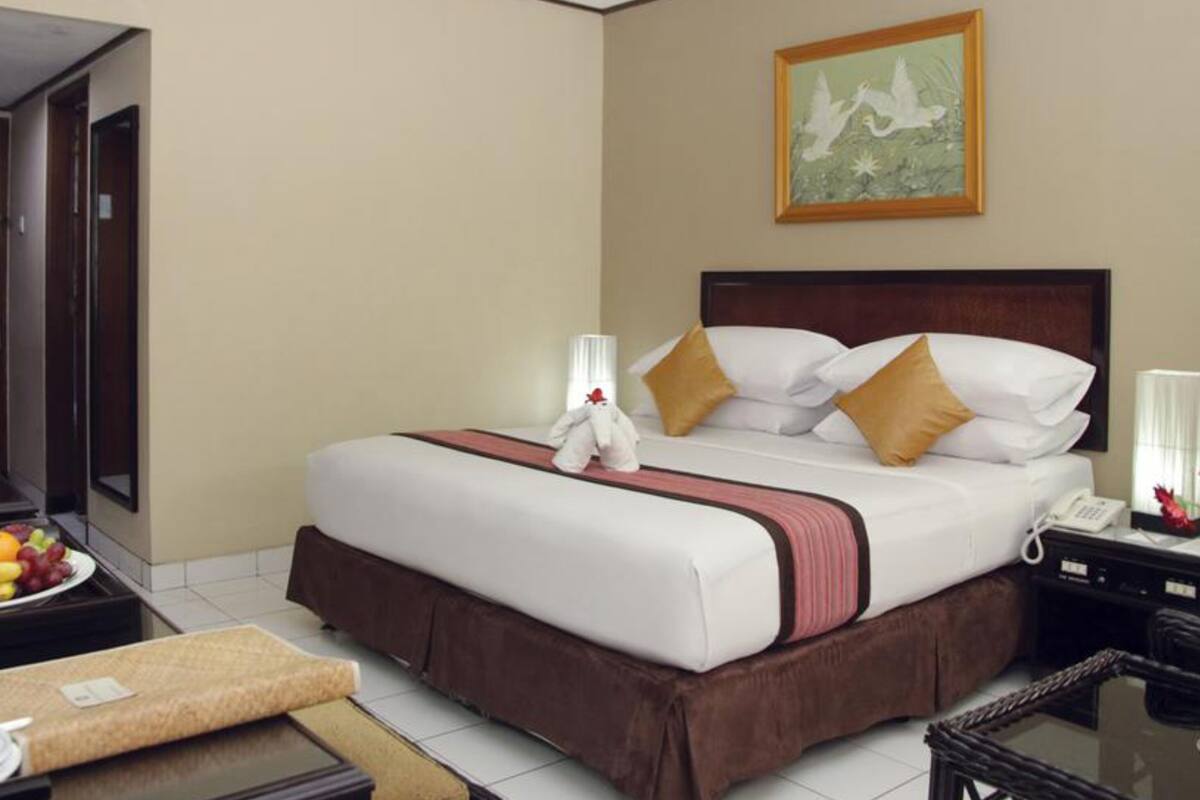 Una habitación de hotel con una cama bien hecha, una cesta de frutas, una mesita de noche con lámpara y un cuadro en la pared.
