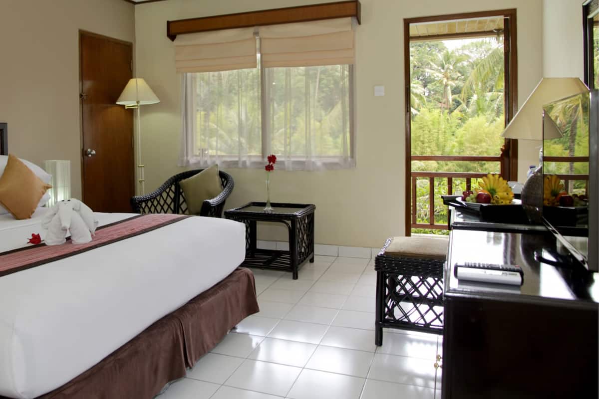 Una habitación de hotel con una cama grande, un área de descanso con una silla y una mesita, un escritorio con una lámpara y una ventana con vista a zonas verdes.