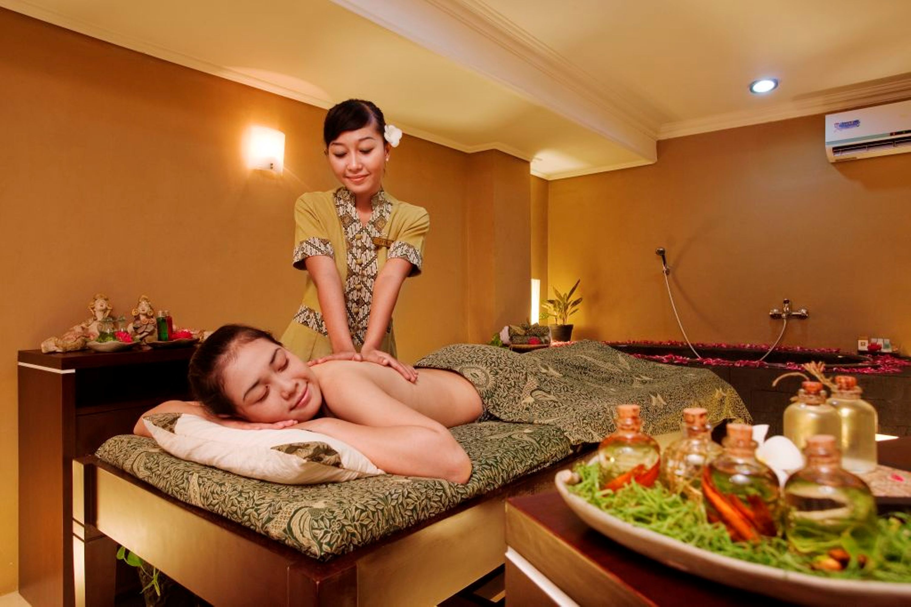 A massage therapy room with a client lying on a massage table, a therapist standing behind, and a tray of oils and herbs on a table.