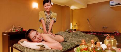 A massage therapy room with a client lying on a massage table, a therapist standing behind, and a tray of oils and herbs on a table.
