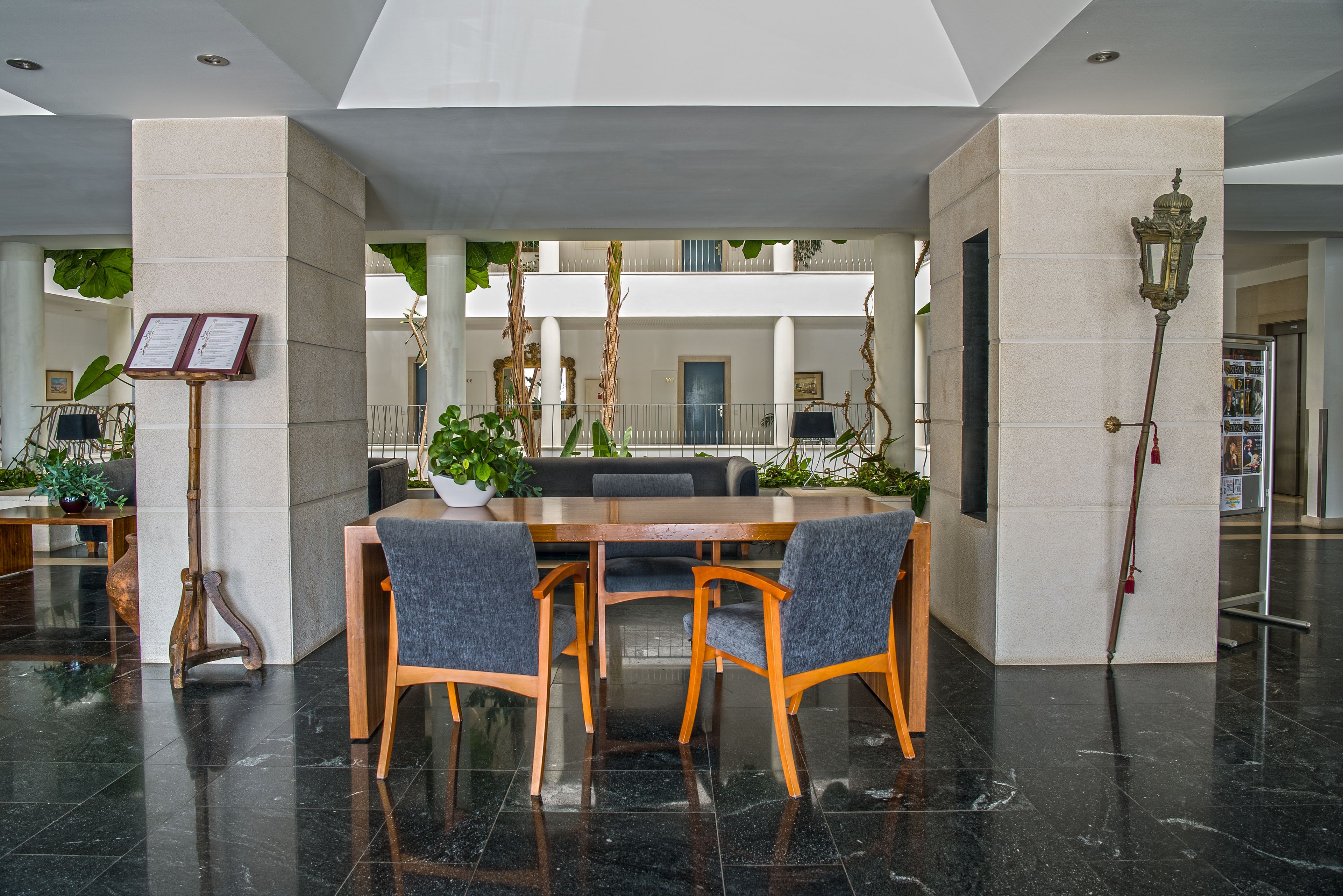 A spacious indoor area with a wooden table and chairs, surrounded by columns and potted plants.