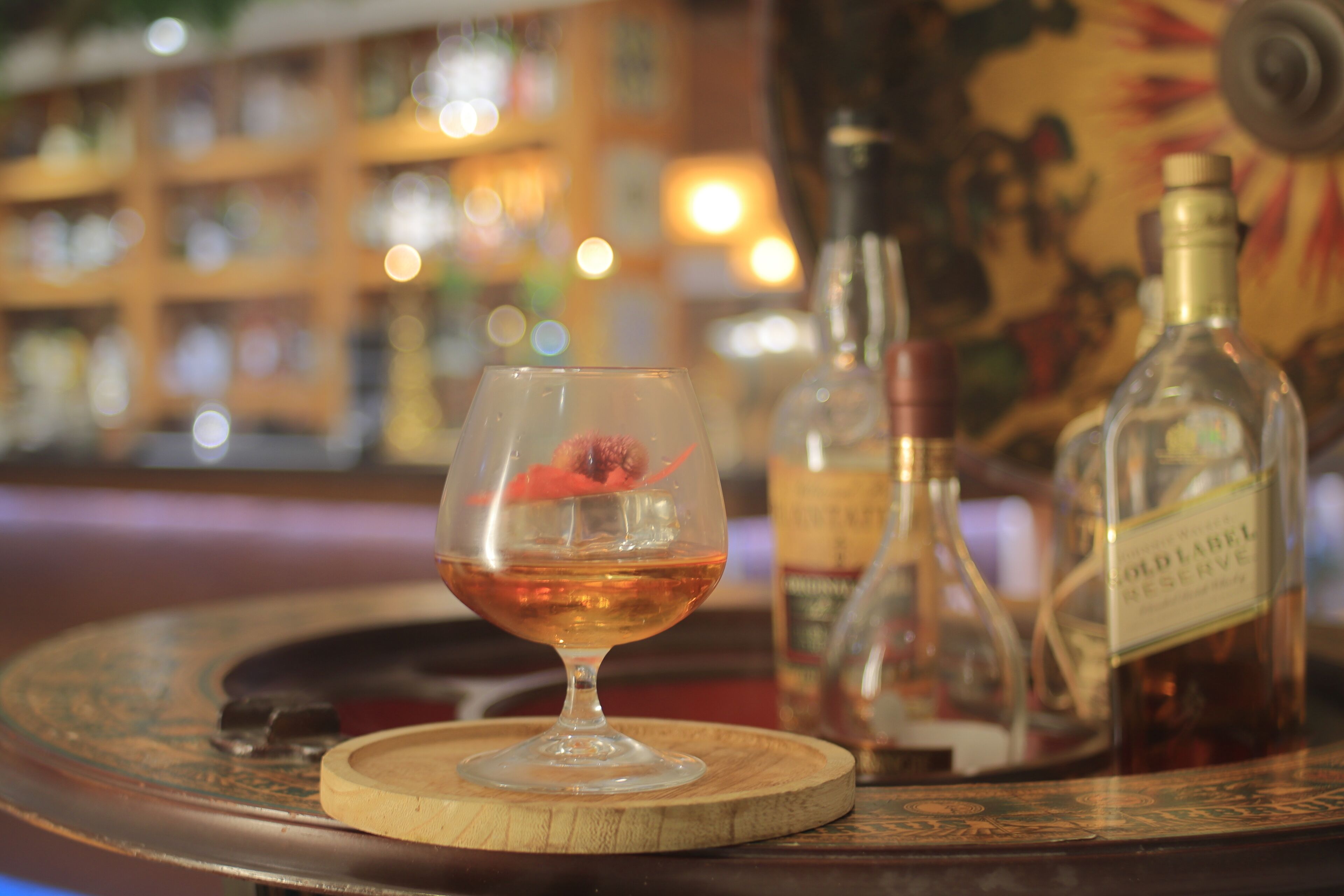 Un bar, avec un verre d’une liqueur ambrée posé sur un sous-verre en bois, entouré de bouteilles de liqueur.