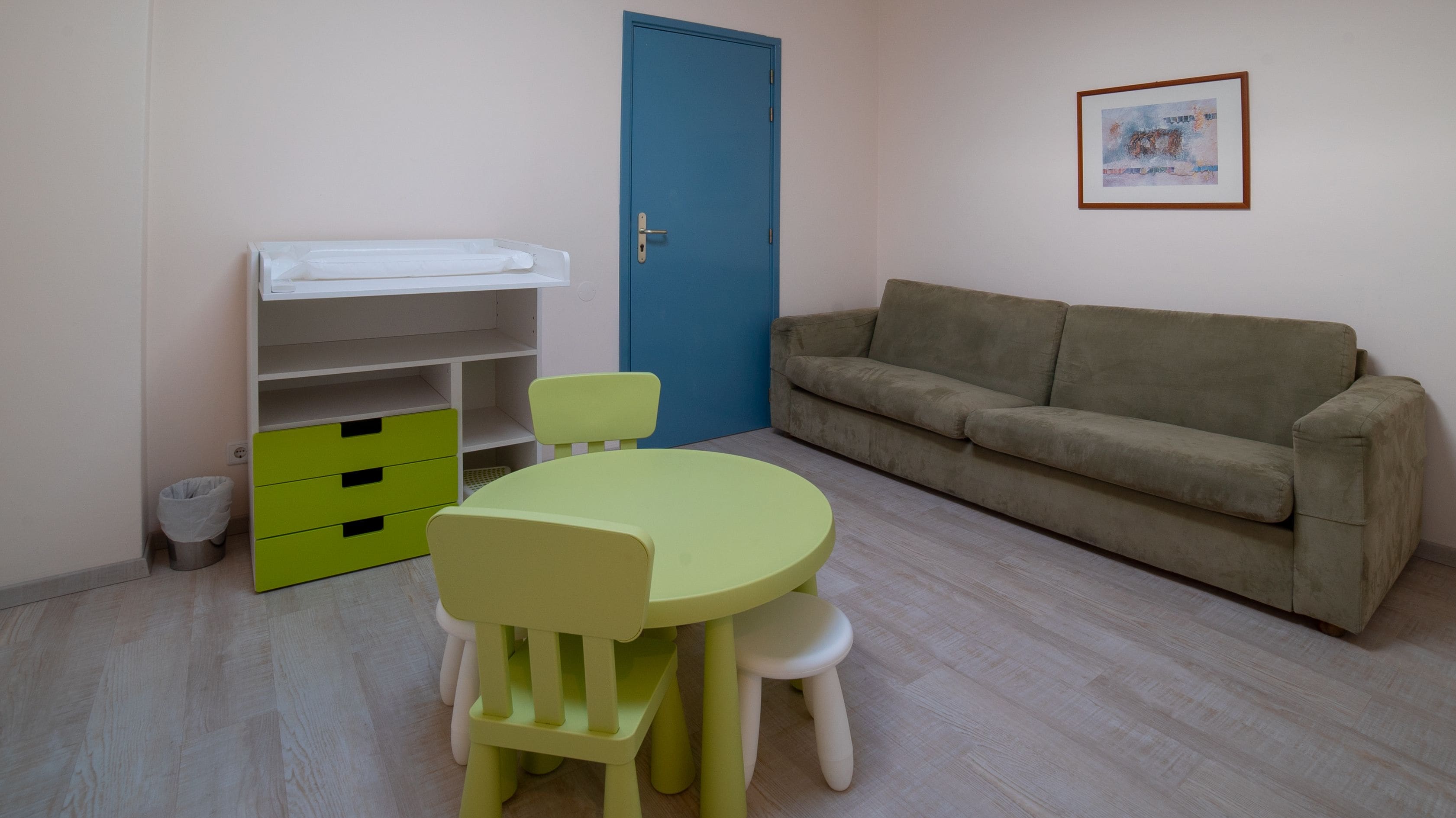 Una habitación con una mesa y sillas verdes, un sofá color beige, una puerta azul y un cuadro en la pared.