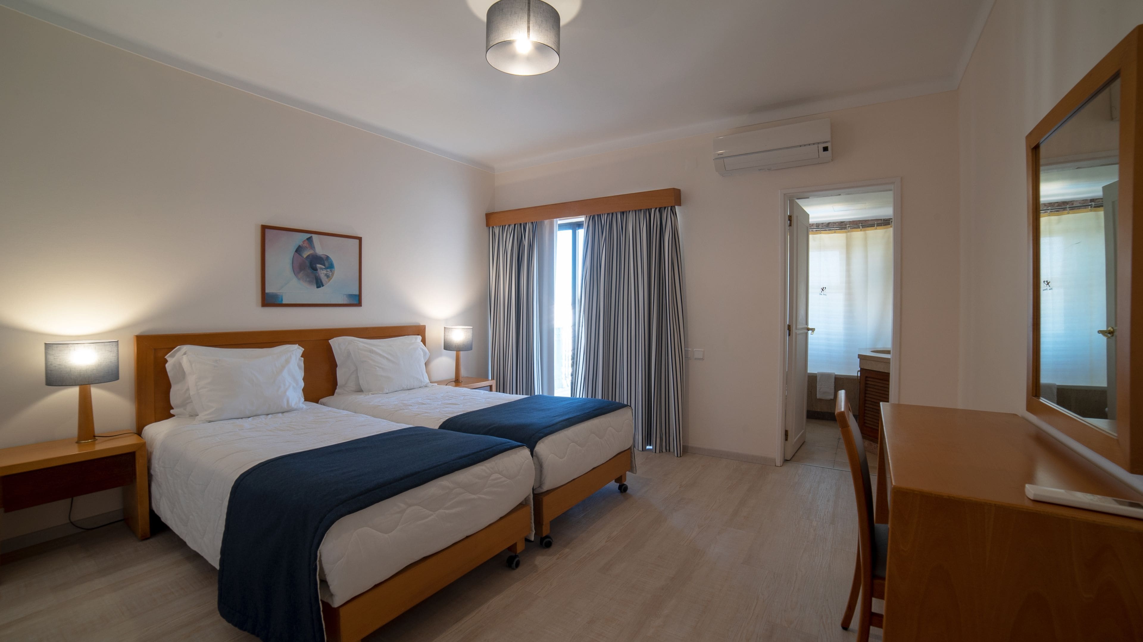 Quarto de hotel com duas camas, uma escrivaninha, uma cadeira, um espelho e banheiro visível por uma porta aberta.