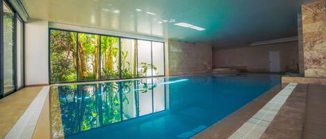 An indoor swimming pool with clear glass walls, a tiled floor, and a view of greenery outside.