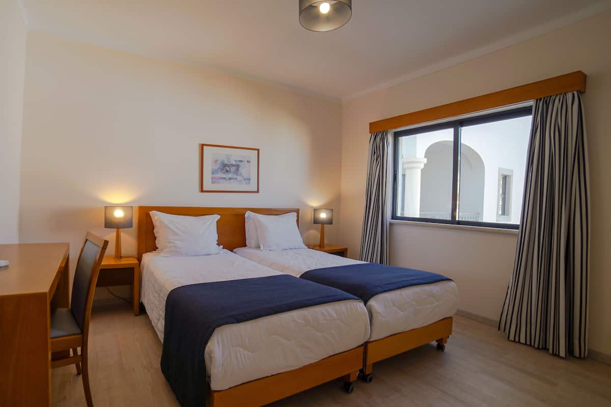 A hotel room with two beds, a desk, a chair, a bedside table, a lamp, a framed picture on the wall, and a window with curtains.