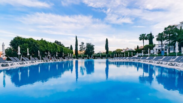 A large, clear swimming pool with lounge chairs and umbrellas along the sides, surrounded by trees and buildings.