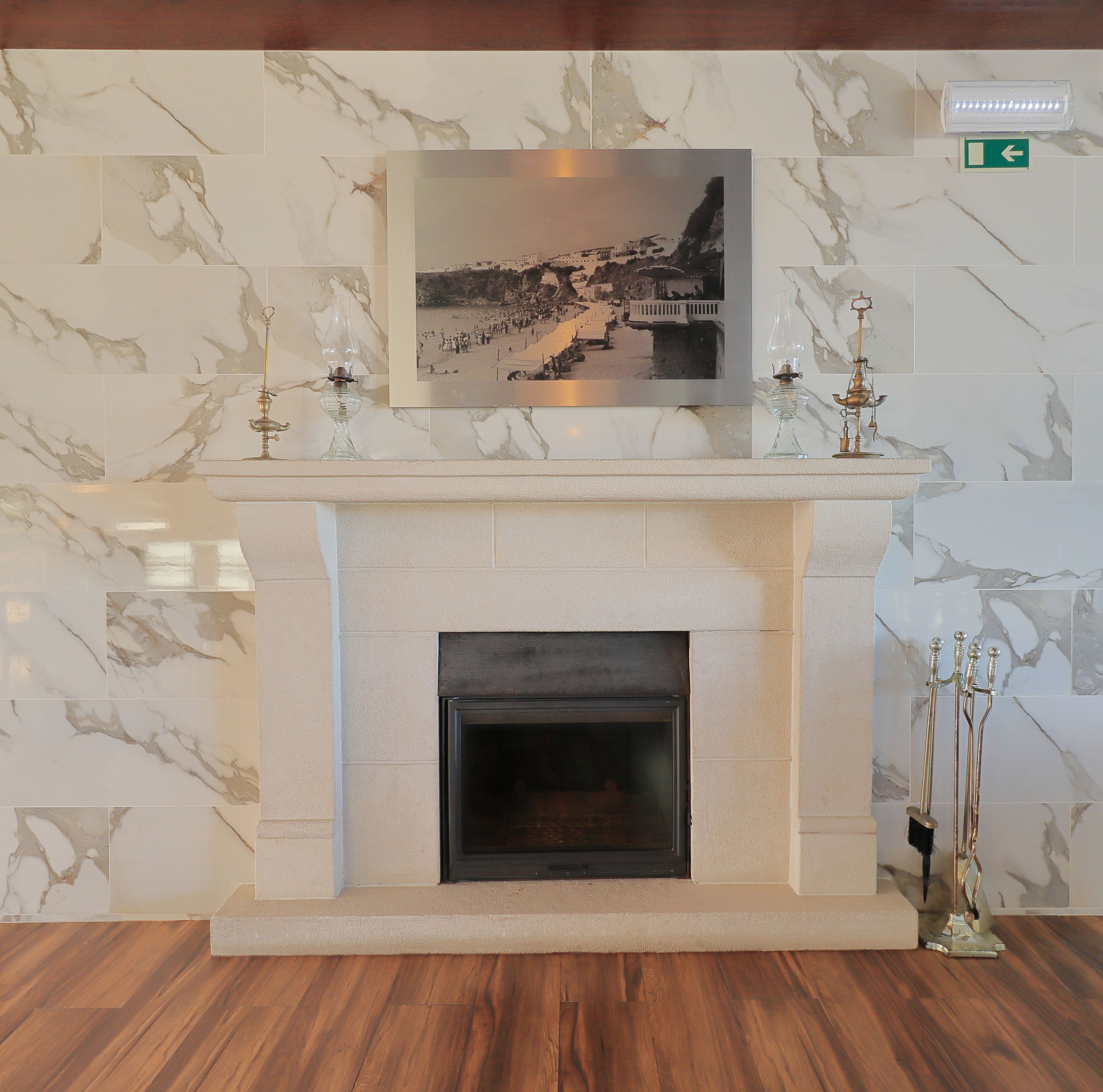 A marble fireplace with a framed photograph above it, a small table with a candle, and a wall-mounted emergency exit sign.