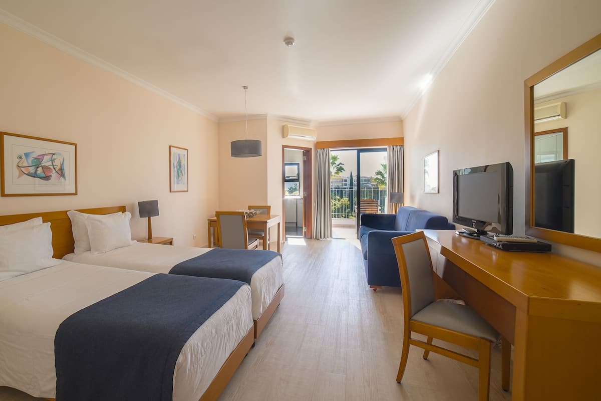 A hotel room with two beds, a desk with a computer, a TV, and a balcony with a view of palm trees.